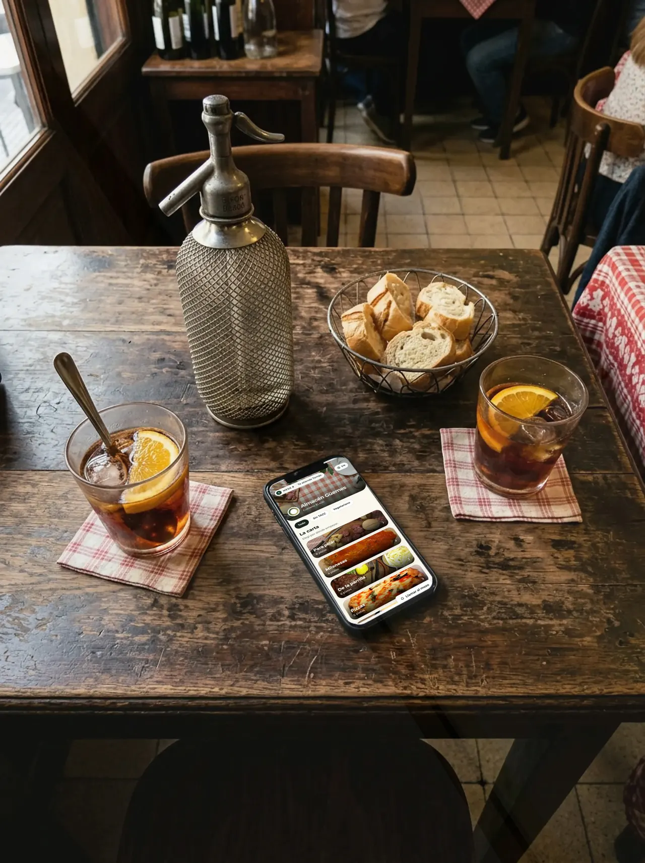 Un teléfono con la carta de Almacén Güemes abierta, apoyado en la mesa de un bar entre dos vermuts y un sifón